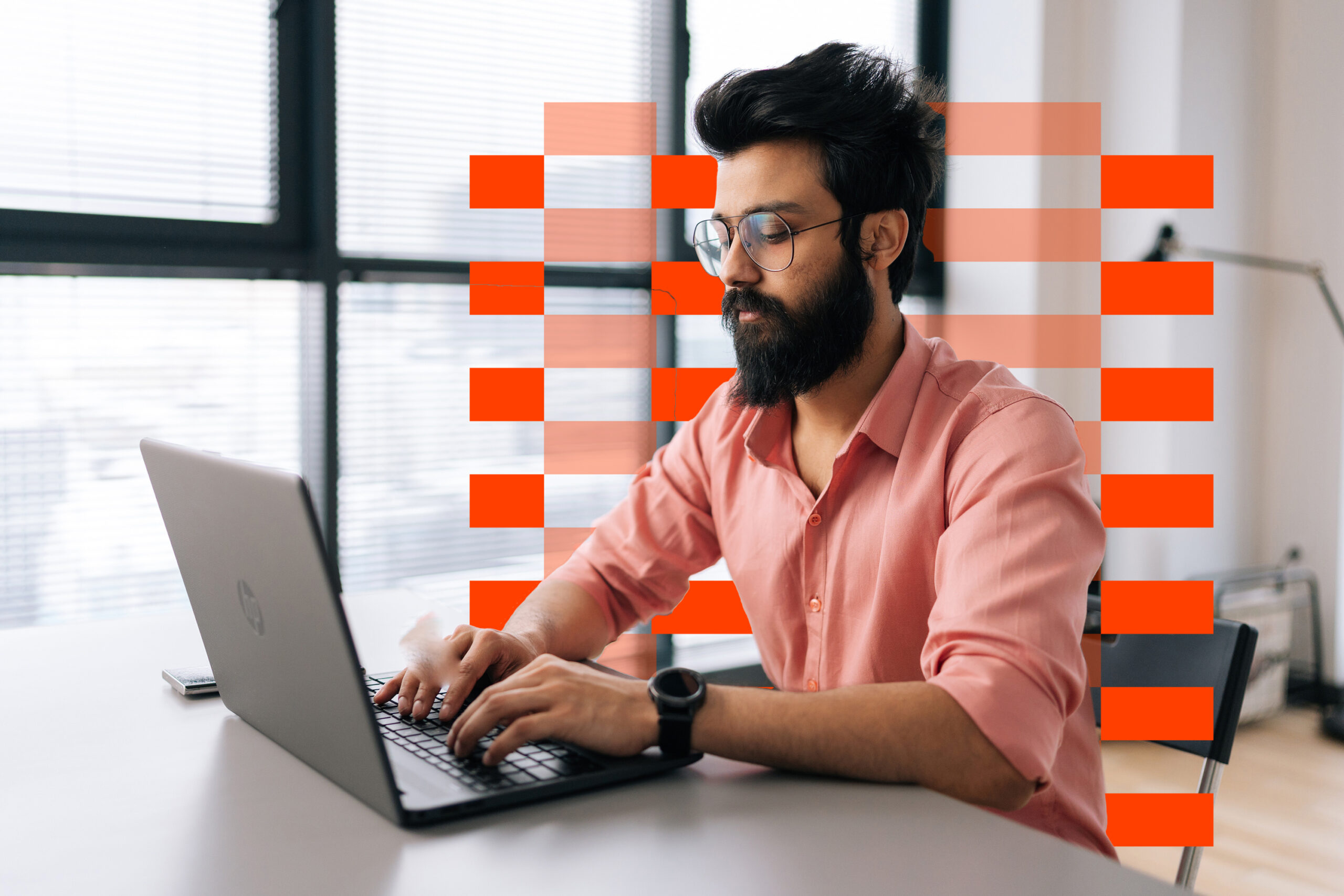 Focused Indian businessman wearing casual clothes working typing on laptop sitting at table in light office room on background of window.